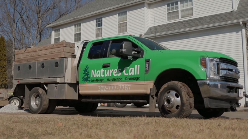 Nature's Call Landscaping branded green Ford pickup truck with stake-body trailer parked on a North Wilmington, Delaware jobsite — Landscape, Hardscape, Drainage signage on the door, phone number 302-777-7767.