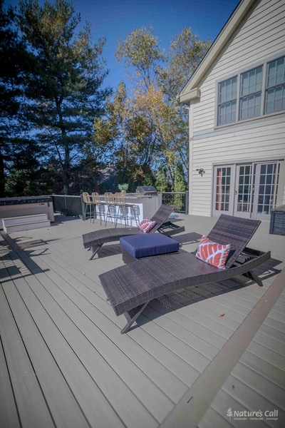 Wide deck-level view of the Chadds Ford back patio in autumn: two wicker chaise lounges in the foreground, the outdoor kitchen bar with wicker stools mid-frame, and the home's tan siding and mature autumn trees in the background