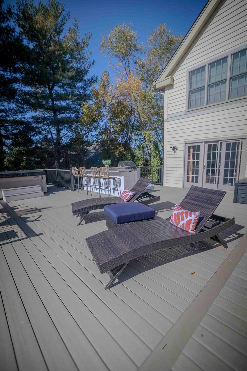 Wide deck-level view of the Chadds Ford back patio in autumn: two wicker chaise lounges in the foreground, the outdoor kitchen bar with wicker stools mid-frame, and the home's tan siding and mature autumn trees in the background