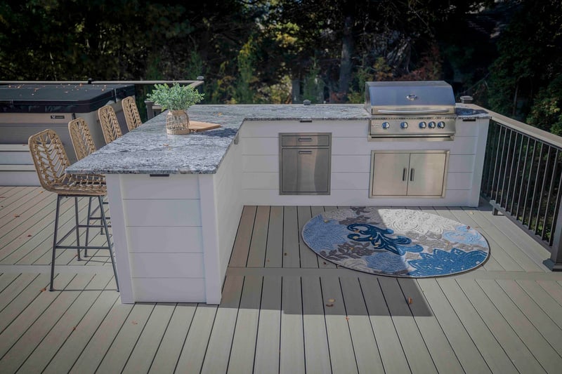 Three-quarter view of the Chadds Ford outdoor kitchen: the granite-topped bar runs along the left, with a side cabinet and stainless gas grill at the center, a circular blue-and-white patterned rug at right, and a single wicker bar stool in profile
