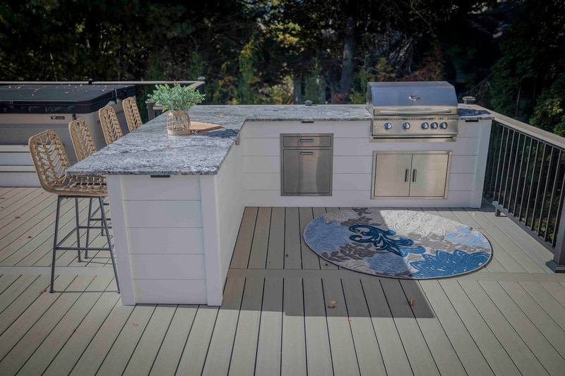 Three-quarter view of the Chadds Ford outdoor kitchen: the granite-topped bar runs along the left, with a side cabinet and stainless gas grill at the center, a circular blue-and-white patterned rug at right, and a single wicker bar stool in profile