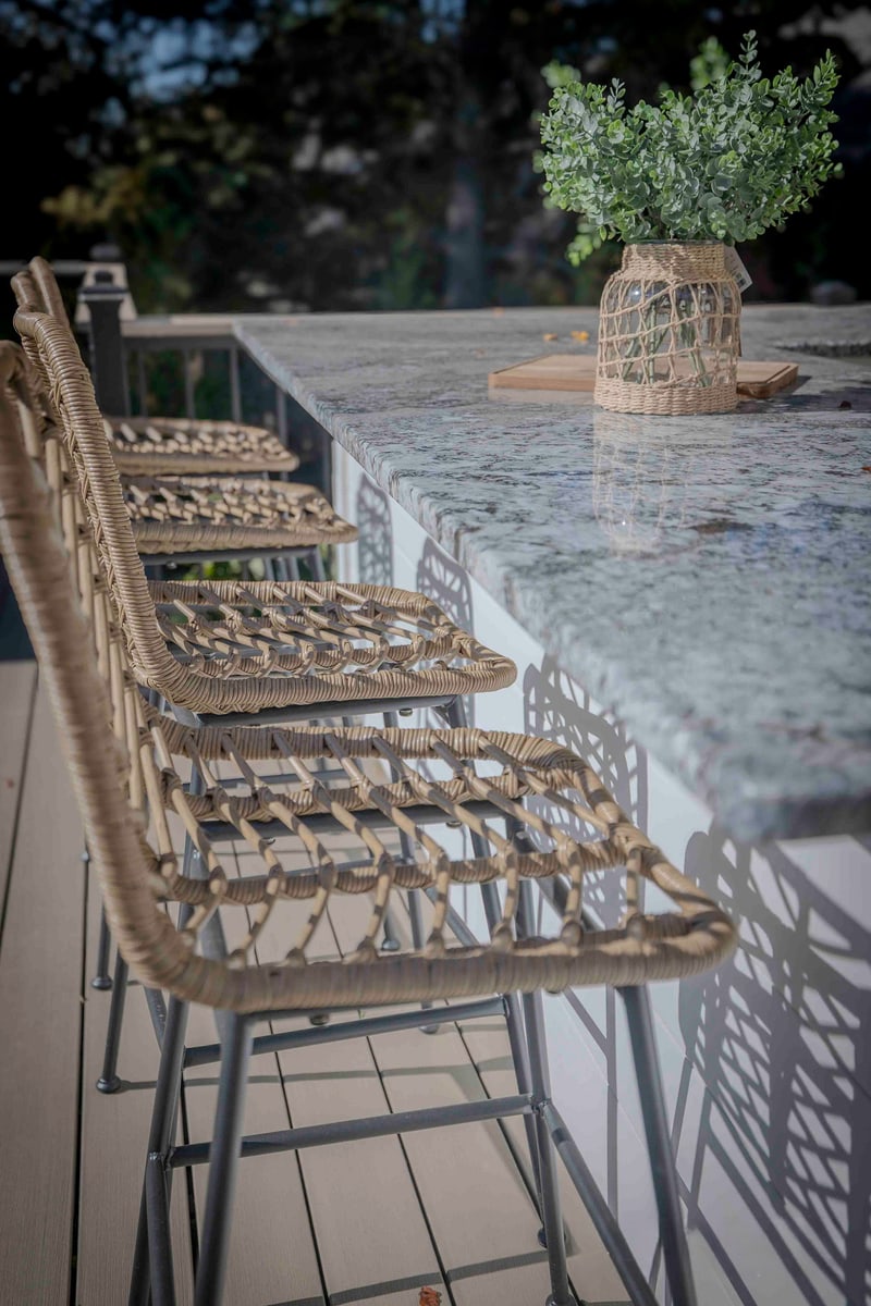 Portrait macro of the Chadds Ford bar stools: four wicker bar-height stools lined up along the granite-topped outdoor kitchen bar, with the cream concrete bar wall and gas grill cabinetry visible behind