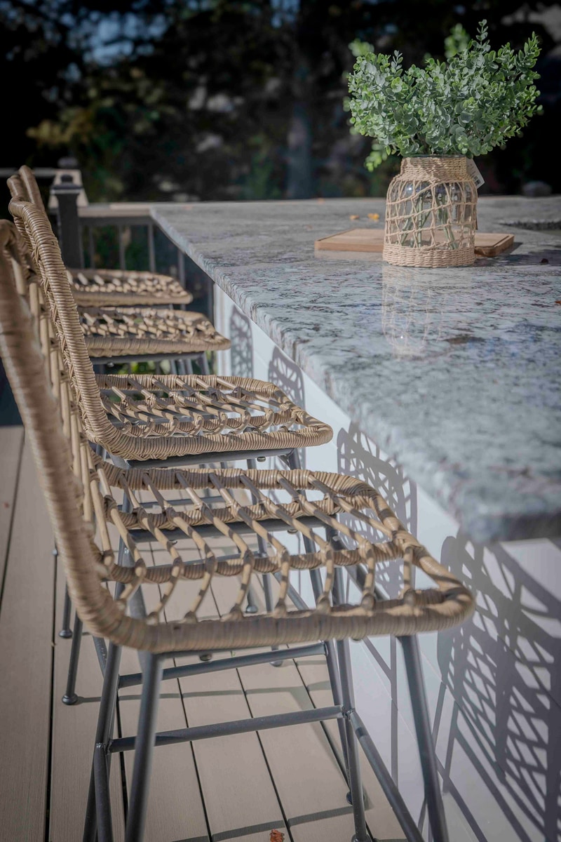 Portrait macro of the Chadds Ford bar stools: four wicker bar-height stools lined up along the granite-topped outdoor kitchen bar, with the cream concrete bar wall and gas grill cabinetry visible behind