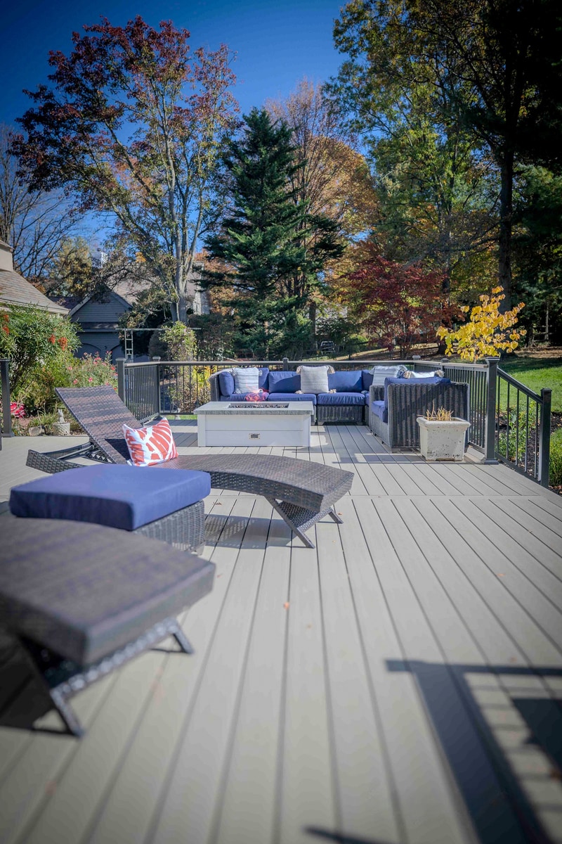 Vivid autumn view of the Chadds Ford back deck with a curved deep blue wicker sectional around a square white propane fire pit, mature trees in red, orange, and yellow fall color filling the background — Nature's Call hardscaping and outdoor living project