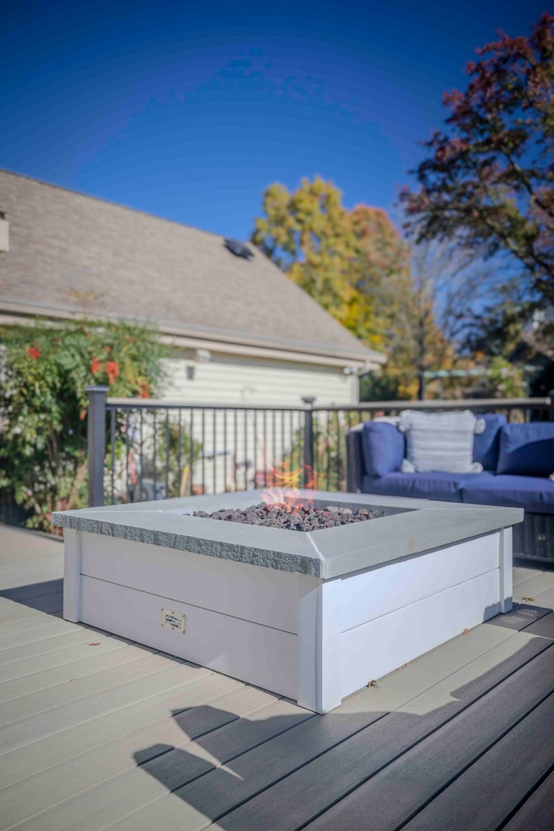 Tight portrait composition of the Chadds Ford fire pit: the square white concrete-topped propane pit fills the lower frame, with the home's tan siding and a red-flowering shrub visible past the black aluminum railing and vivid autumn trees overhead
