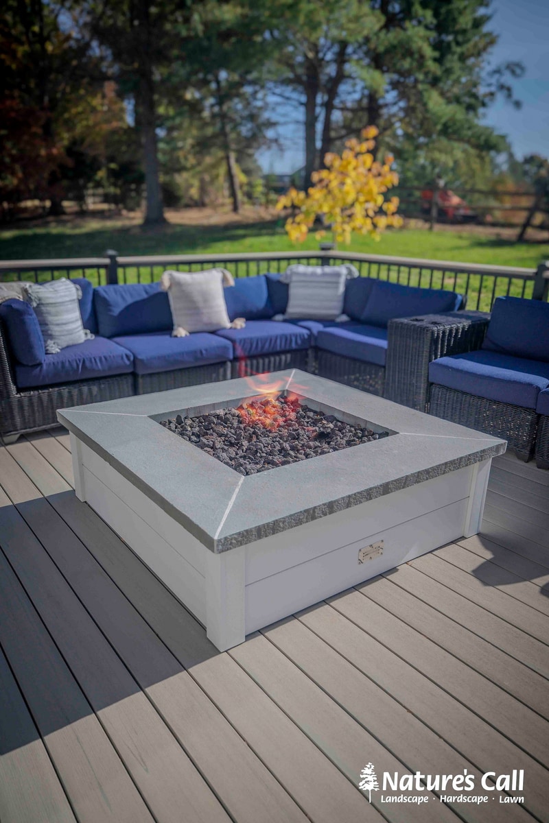 Vertical cinematic composition of the Chadds Ford fire pit: a square white concrete-topped propane fire pit with lit lava rocks centered on the gray composite deck, the curved deep blue wicker sectional behind, and a single yellow-foliage autumn tree silhouetted against the sky