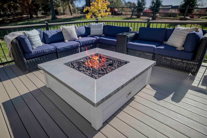 Magazine-style close-up of the lit propane fire pit on the Chadds Ford composite deck: lava-rock flames burning in the white concrete-topped square pit, a deep blue wicker sectional with cream pillows wrapping three sides, autumn trees and a distant red barn beyond the black aluminum railing