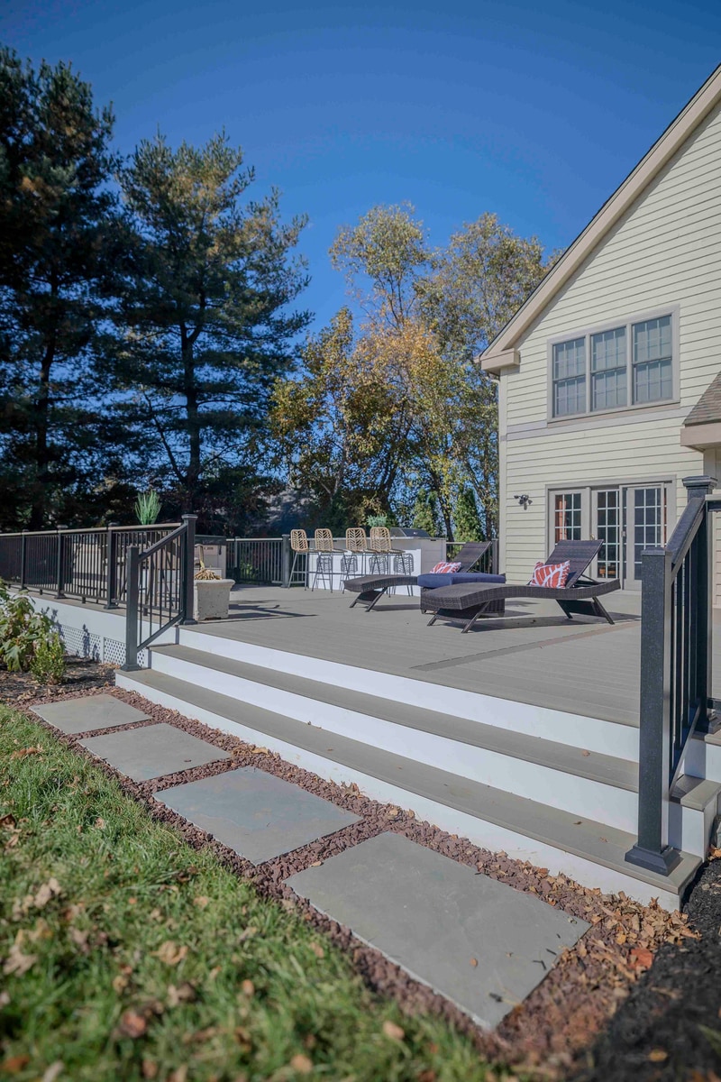 Portrait view of the bluestone stepping stones leading from the lawn up to the Chadds Ford composite deck: irregular bluestone treads set into a leaf-strewn mulch bed, with the deck edge and a wicker bar stool visible above