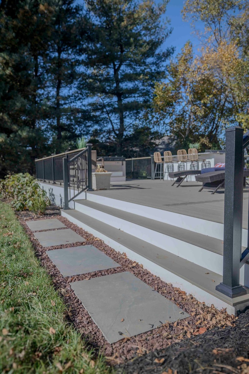Portrait view looking up the bluestone steps to the Chadds Ford deck: irregular bluestone treads set into a mulch and leaf bed lead the eye up to the white riser of the deck step, with wicker bar stools and a sunlit chaise visible above