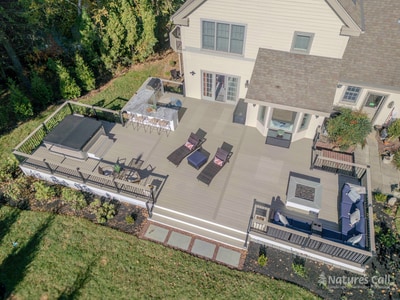 Aerial three-quarter view of the Chadds Ford back deck showing the entire project in one frame: outdoor kitchen with bar seating at the top, recessed hot tub on the left, two wicker chaise lounges in the center, and the curved deep blue wicker sectional surrounding the square white propane fire pit at the bottom right, all set against the home's tan siding and the autumn tree line
