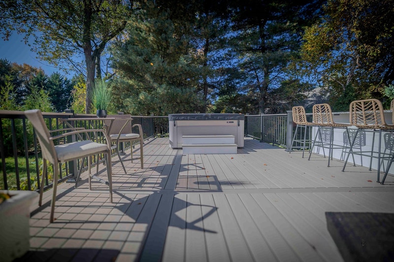 Wide deck-level view of the Chadds Ford hot tub zone: the recessed hot tub at center frame ringed by a small chair seating area on the left and the outdoor kitchen bar with wicker stools on the right, all on the gray composite deck under autumn trees