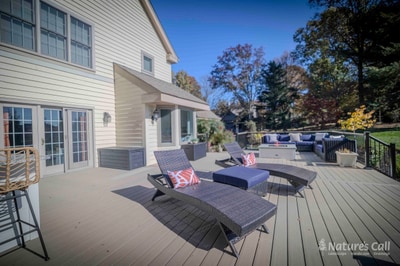 Wide deck-level view across the Chadds Ford composite deck: two wicker chaise lounges with patterned cream-and-orange pillows in the foreground, the curved deep blue sectional and square fire pit visible in the middle-right, and the home's tan siding wrapping the left edge under a clear blue autumn sky