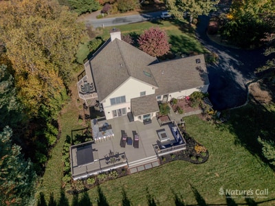 Aerial three-quarter view of the front of the Chadds Ford home in autumn: the two-story tan-siding house with stone chimney sits among mature trees in peak red, yellow, and burgundy fall color, with the back patio just visible beyond the roofline