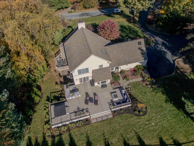 Aerial three-quarter view of the front of the Chadds Ford home in autumn: the two-story tan-siding house with stone chimney sits among mature trees in peak red, yellow, and burgundy fall color, with the back patio just visible beyond the roofline