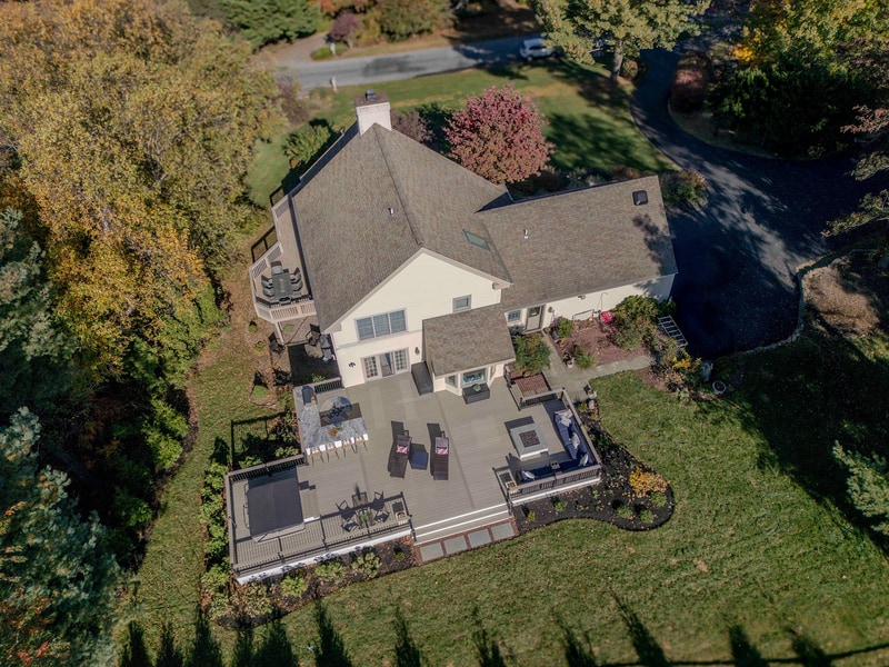 Aerial three-quarter view of the front of the Chadds Ford home in autumn: the two-story tan-siding house with stone chimney sits among mature trees in peak red, yellow, and burgundy fall color, with the back patio just visible beyond the roofline