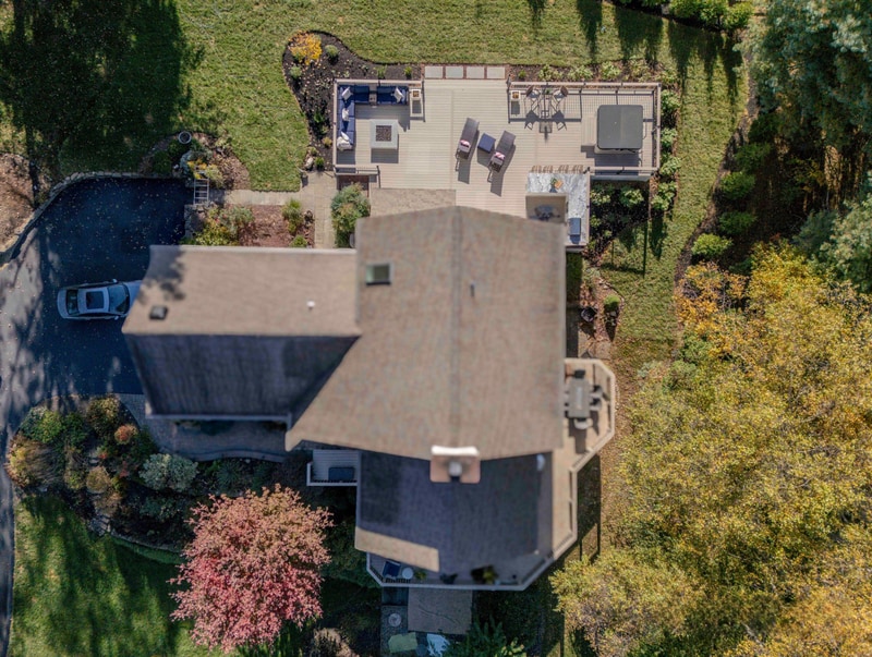 Sun-drenched ground-level view of the Chadds Ford fire pit zone: the square white propane fire pit at center frame, ringed by a curved deep blue wicker sectional with cream pillows on the left, two wicker chaise lounges on the right, and a neighbor's home visible in the distance