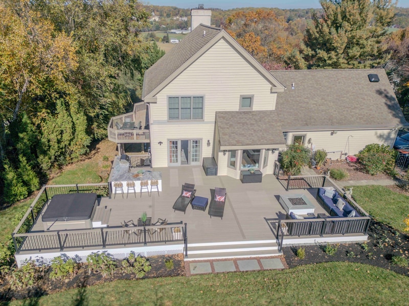 Aerial three-quarter view of the full Chadds Ford back deck embedded in autumn landscape: the multi-zone composite deck with bar seating, two wicker chaise lounges, recessed hot tub, and curved sectional fire pit lounge laid out behind the tan-siding home, with mature autumn trees and rolling hills filling the background