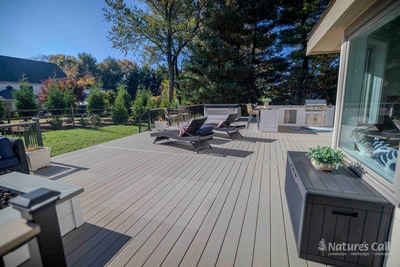 Wide ground-level view of the Chadds Ford back deck looking toward the home: a wicker chaise lounge with a red-pattern pillow in the foreground, the outdoor kitchen with bar seating mid-frame, and the deep blue sectional and french doors of the home visible in the distance