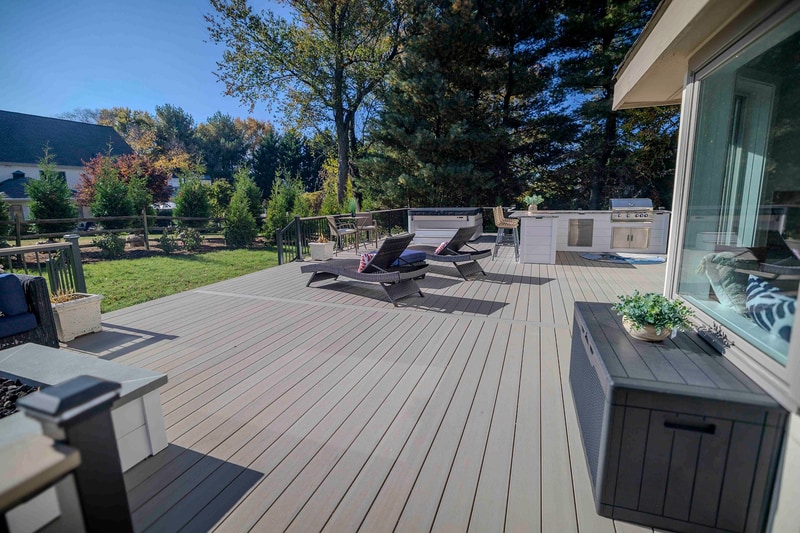 Wide ground-level view of the Chadds Ford back deck looking toward the home: a wicker chaise lounge with a red-pattern pillow in the foreground, the outdoor kitchen with bar seating mid-frame, and the deep blue sectional and french doors of the home visible in the distance