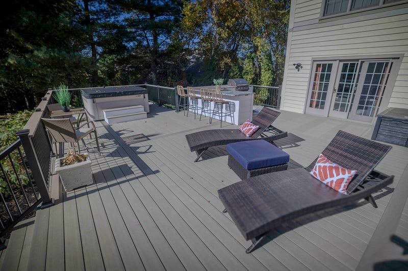 Deck-level view of the Chadds Ford back patio looking toward the home's french-door entry: a wicker chaise lounge and ottoman in the foreground, a chair grouping with painted-orange-and-cream pillows mid-frame, and the outdoor kitchen against the left edge