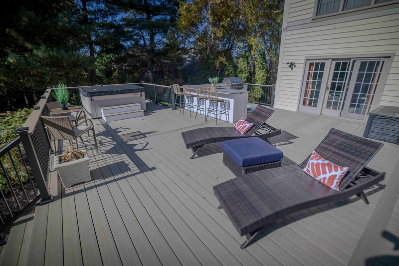 Deck-level view of the Chadds Ford back patio looking toward the home's french-door entry: a wicker chaise lounge and ottoman in the foreground, a chair grouping with painted-orange-and-cream pillows mid-frame, and the outdoor kitchen against the left edge