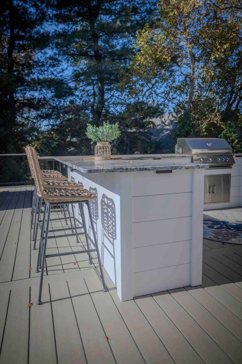 Sun-streaked portrait of the Chadds Ford outdoor kitchen: four wicker bar-height stools line the granite-topped bar, with the gas grill and storage cabinetry visible at the right and a small potted plant on the counter