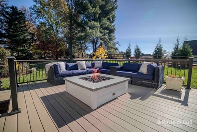 Wide ground-level view of a Chadds Ford deck zone with a U-shaped deep blue wicker sectional surrounding the white square fire pit, autumn trees and lawn visible past the black aluminum railing