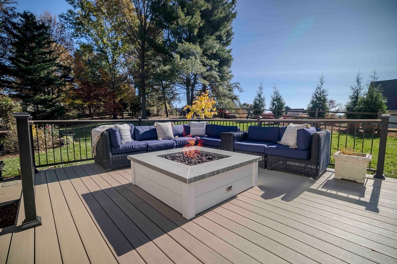 Wide ground-level view of a Chadds Ford deck zone with a U-shaped deep blue wicker sectional surrounding the white square fire pit, autumn trees and lawn visible past the black aluminum railing