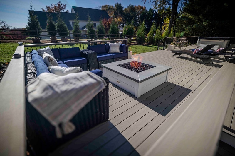 Wide cinematic ground-level view of the Chadds Ford back deck on a clear autumn day: a square white propane fire pit at center frame, ringed by a curved deep blue wicker sectional with cream pillows on the left, two wicker chaise lounges on the right, and the home's tan siding visible at the far edge