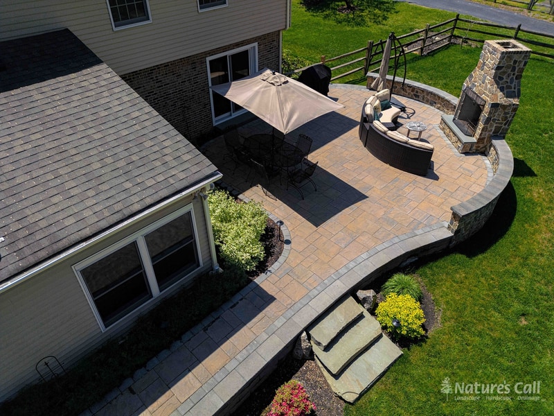 Aerial view of a Garnet Valley Pennsylvania back patio: a curved paver platform surrounds an in-ground hot tub and full-height fieldstone outdoor fireplace, with a tan umbrella shading the dining area — Nature's Call landscaping and hardscaping project