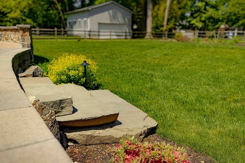 Corner of the low fieldstone retaining wall meeting the paver walkway in Garnet Valley, with a path light and single yellow-foliage shrub in the foreground and the lawn extending toward a distant garage