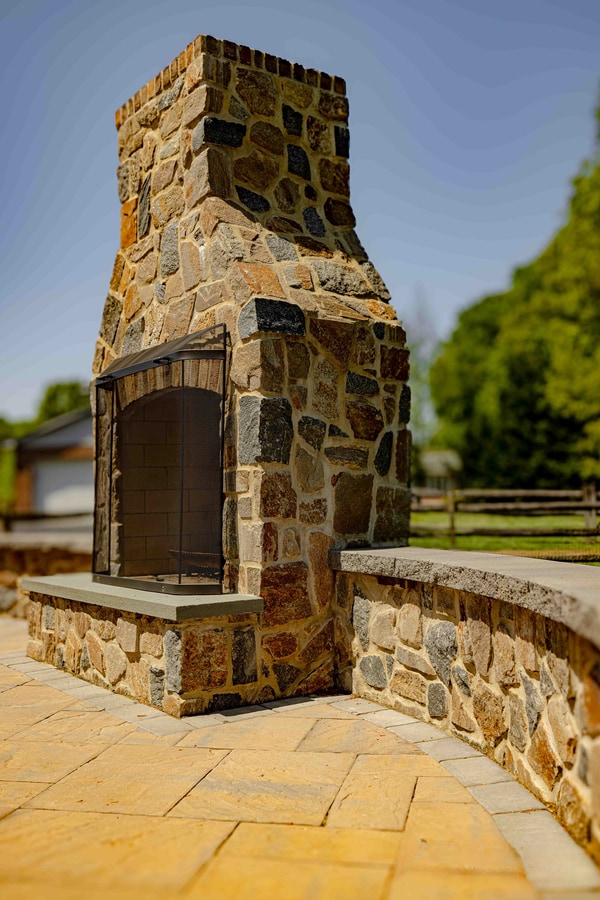 Hand-cut fieldstone outdoor fireplace with stone hearth and capstone bench return on a Garnet Valley paver patio — Nature's Call Landscaping