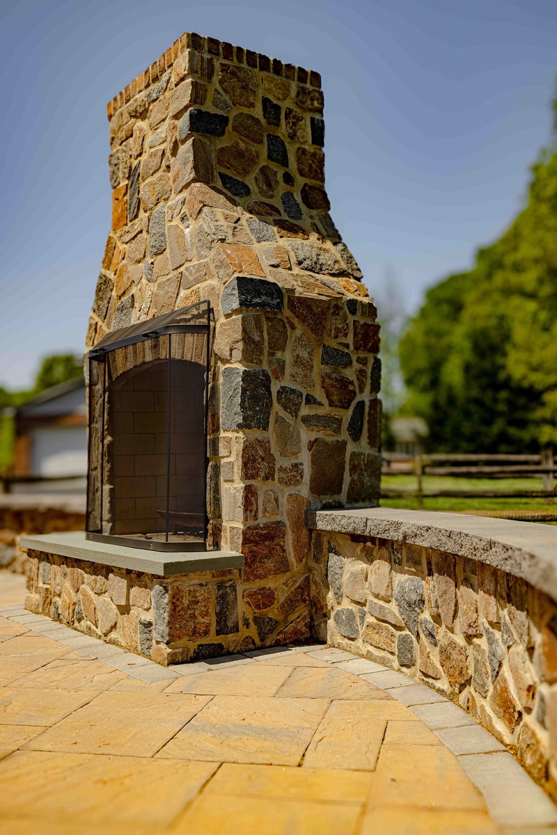 Looking up at the full-height fieldstone outdoor fireplace at the Garnet Valley project, brick crown at the top of the chimney and the patio's stone perimeter wall visible at the base