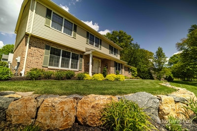 Three-quarter front view of the Garnet Valley home from a low angle, with large fieldstone boulders and ribbons of daylilies anchoring the front planting bed in the foreground and a clear blue sky overhead