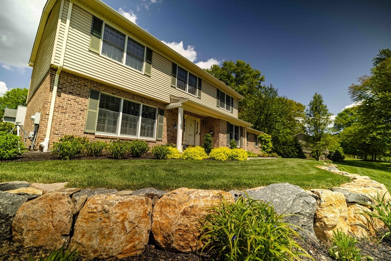 Three-quarter front view of the Garnet Valley home from a low angle, with large fieldstone boulders and ribbons of daylilies anchoring the front planting bed in the foreground and a clear blue sky overhead