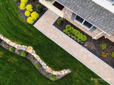 Drone view straight down on the Garnet Valley front walkway: a charcoal-bordered tan paver path runs between mulched beds of yellow-foliage shrubs, with a curved fieldstone retaining wall along the lawn edge