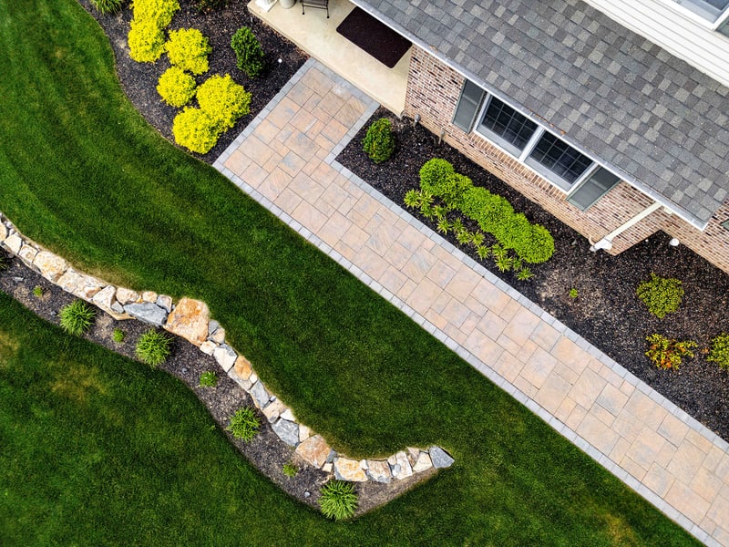 Drone view straight down on the Garnet Valley front walkway: a charcoal-bordered tan paver path runs between mulched beds of yellow-foliage shrubs, with a curved fieldstone retaining wall along the lawn edge