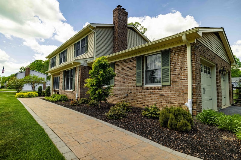 Side view of the Garnet Valley home showing the brick chimney, with the paver walkway curving from the driveway around the house past a mulched bed of mounded shrubs