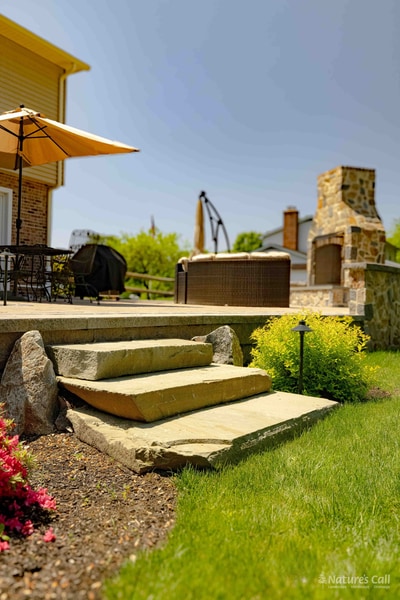 Hand-laid flagstone steps rising from the lawn to the Garnet Valley back patio, with a tan umbrella, hot tub, and fieldstone fireplace visible above and a yellow-foliage shrub at right