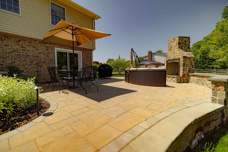 Ground-level view of the Garnet Valley back patio from the lawn corner, with the brick home on the right, the full-height fieldstone fireplace mid-right, and a tan umbrella shading the dining area