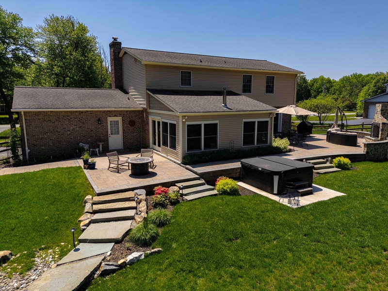 Elevated view of the rear of the Garnet Valley home, showing the multi-level paver patio with dining set, the in-ground hot tub, and stone steps leading down to the lawn