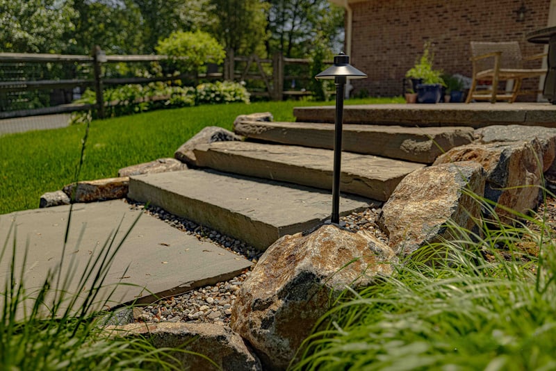 Hand-laid flagstone garden steps cutting through a planted bed at the Garnet Valley project, with a bronze low-voltage path light beside the steps and ornamental grasses framing the foreground