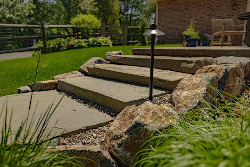 Hand-laid flagstone garden steps cutting through a planted bed at the Garnet Valley project, with a bronze low-voltage path light beside the steps and ornamental grasses framing the foreground