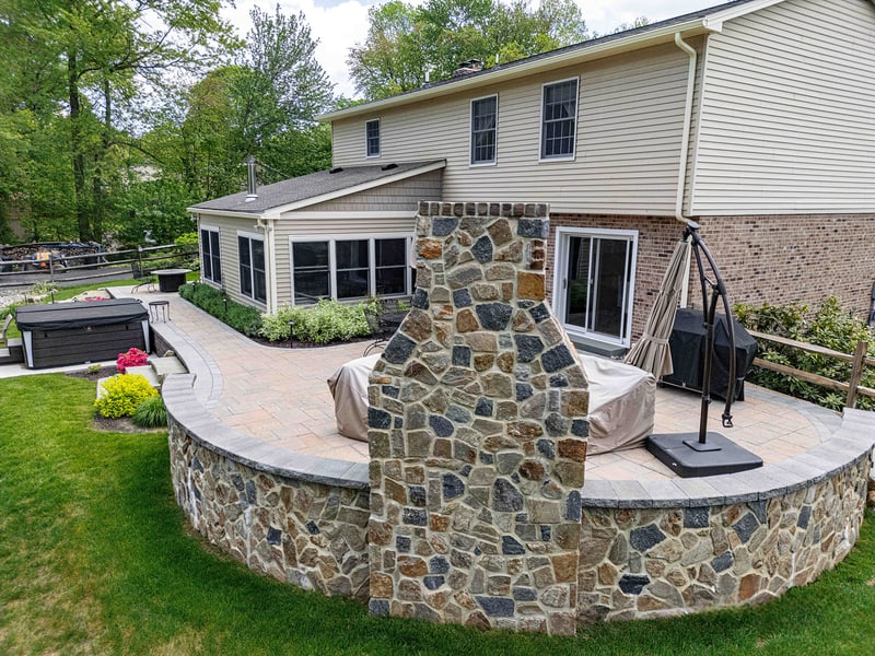 Ground-level view of the Garnet Valley back patio: a full-height fieldstone outdoor fireplace anchors the center, with the in-ground hot tub on the right and a charcoal grill against the brick side of the home