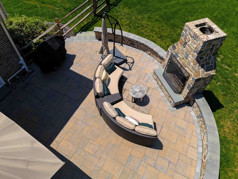 Top-down aerial of the Garnet Valley back patio: a curved seating area with cushioned chairs faces a stone-edged round fire pit, with the full-height fieldstone outdoor fireplace at the right edge of the paver platform