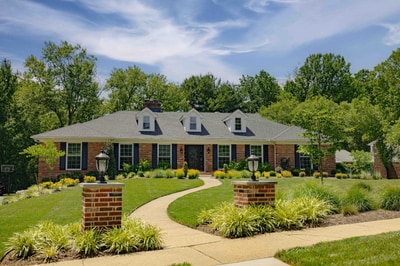 Front view of the Greenville Delaware estate after Nature's Call landscape and hardscape work: brick lamp pillars flanking a stone walkway, layered four-season plantings, and a manicured front lawn.
