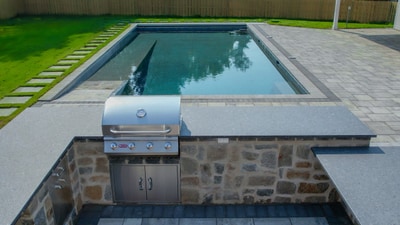 Rectangular pool with stacked stone outdoor kitchen featuring a built-in grill, adjacent paver patio, and manicured lawn.
