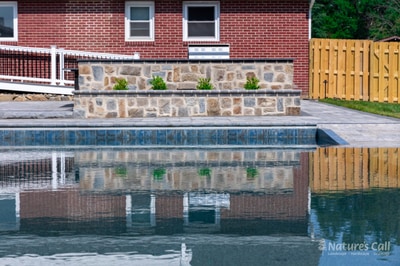 Modern pool with stacked stone retaining wall, foundation plantings, and herringbone brick deck adjacent to brick home.