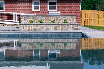 Modern pool with stacked stone retaining wall, foundation plantings, and herringbone brick deck adjacent to brick home.