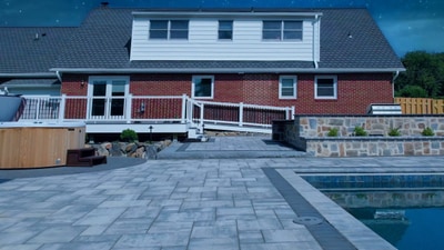 Large paver patio with swimming pool, stacked stone retaining wall, white deck railing, and mature landscape plantings.